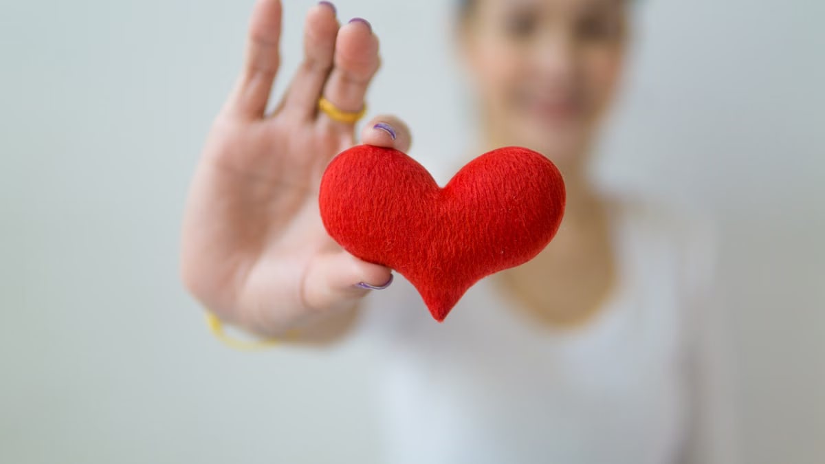 Unrecognizable blurred female standing on white background with outstretched arm while demonstrating red decorative heart in light studio in daytime